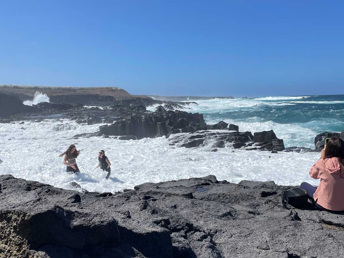An der Küste fotografiert eine Frau das stürmische Meer, während zwei weitere Frauen von hohen Wellen überrascht und beinahe ins Meer gespült werden.